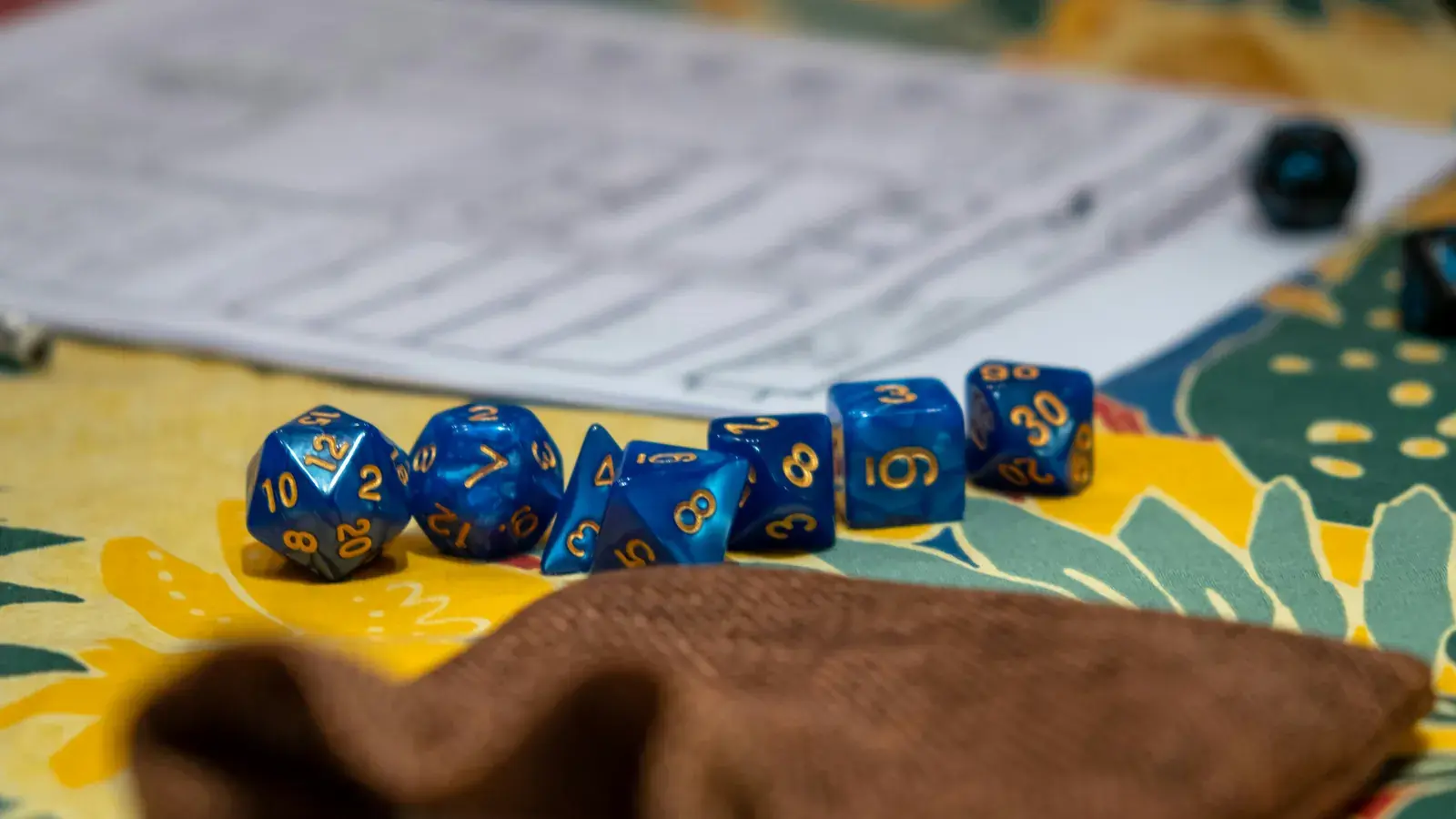 A blue and gold polyhedral dice set lined up in front of a printed character sheet, with a brown dice bag on a patterned tablecloth
